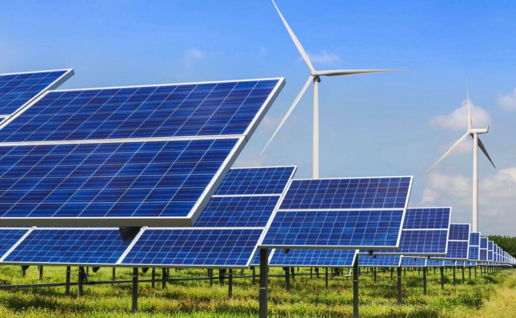 Rows of solar panels stretch into the distance at a solar farm, with wind turbines in the background