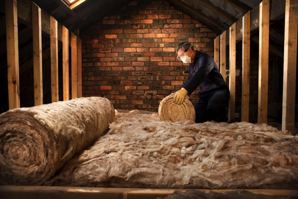 A man wearing a mask and goggles rolls out a layer of insulating material inside a brick and timber loft