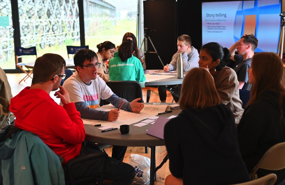 Young people sit in groups around tables discussing ideas for creating a film